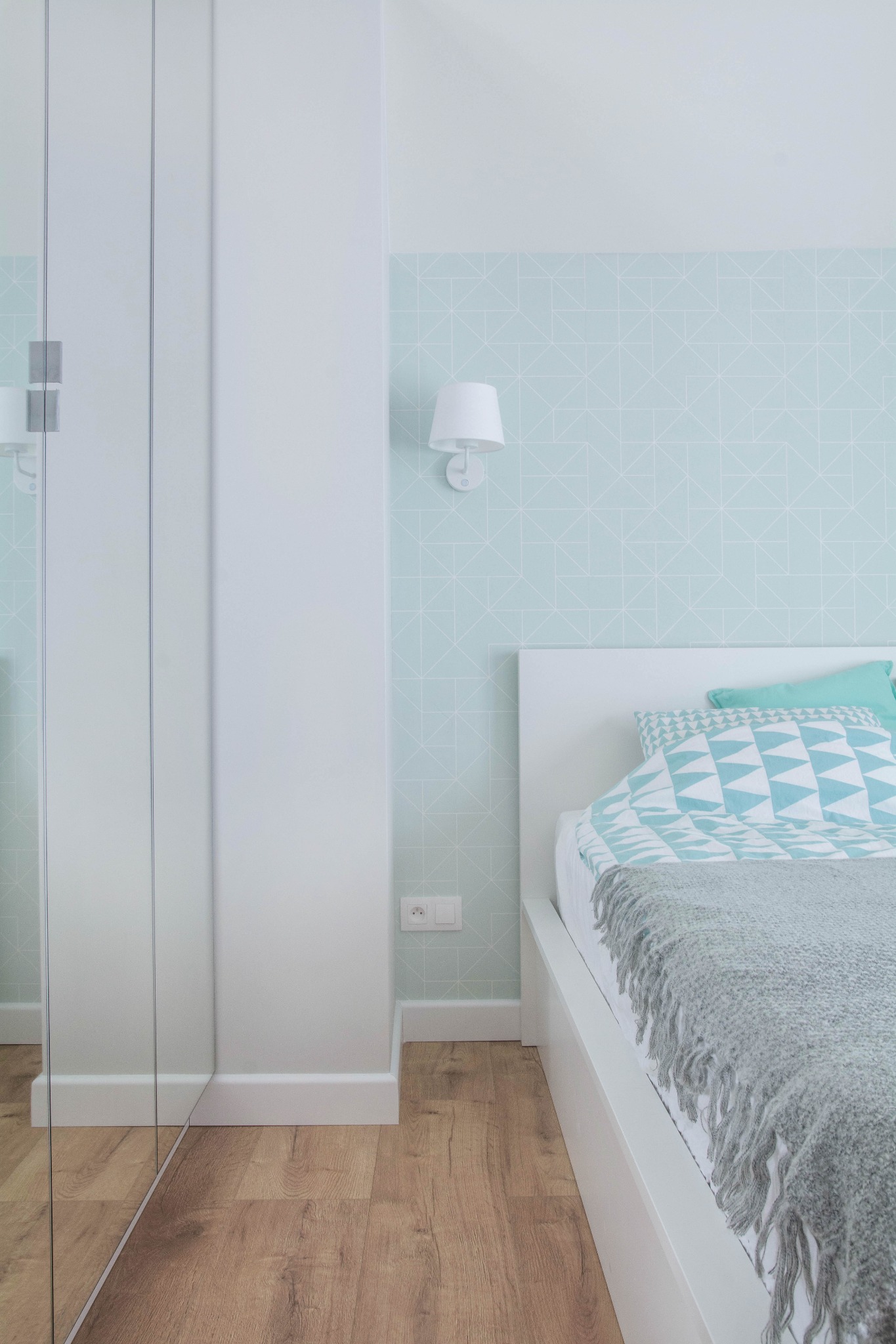 Minimalistyczna sypialnia z białym łóżkiem, wzorzystą pościelą w odcieniach błękitu i szarą narzutą z frędzlami, na ścianie geometryczna tapeta i biały kinkiet, obok szafa z lustrzanymi drzwiami.