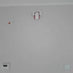 Centrala alarmowa z dwoma czujnikami ruchu zamontowana na szarej ścianie, widoczne kołki montażowe, regulator temperatury i włącznik światła.