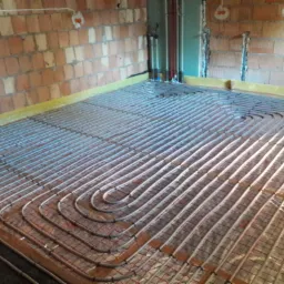 Rozłożone rury ogrzewania podłogowego na folii z siatką, widoczne fragmenty ściany z czerwonej cegły i pionowe rury instalacji w surowym stanie.