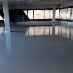 Świeżo wylana, gładka wylewka anhydrytowa w surowym wnętrzu budynku z betonowymi kolumnami i zabezpieczonymi oknami, odbijająca światło w mokrej powierzchni.