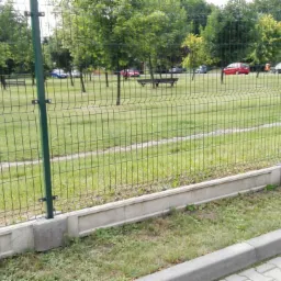 Zielona siatka ogrodzeniowa panelowa z podmurówką betonową oddzielająca trawnik z drzewami od chodnika z kostki brukowej.