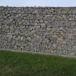 Gabionowa konstrukcja ogrodzeniowa wypełniona szarym kamieniem, widoczna trawa u podstawy, pochmurne niebo w tle.