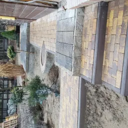 Ułożona kostka brukowa w odcieniach żółci i szarości prowadząca do schodów; w tle widoczne drzewa i elementy drewnianej konstrukcji domu.