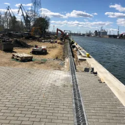 Budowa nabrzeża portowego: ułożona kostka brukowa, zamontowany system odwadniający, w tle pracujące koparki i dźwigi portowe.
