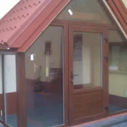 Zadaszone wejście do budynku z brązowymi drzwiami i przeszklonymi ścianami bocznymi, zwieńczone czerwoną blachą trapezową, widoczne z zewnątrz.