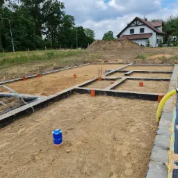 Fundamenty domu jednorodzinnego w trakcie budowy, z widocznymi rurami kanalizacyjnymi i elementami szalunku, na tle domu i hałdy piasku.