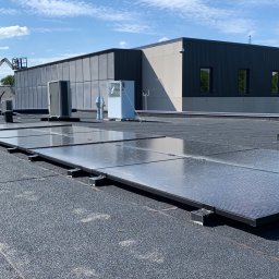 ART-BUD - Instalacja paneli słonecznych na płaskim dachu budynku przemysłowego, widoczne panele, klimatyzatory i pracownicy w oddali, błękitne niebo.