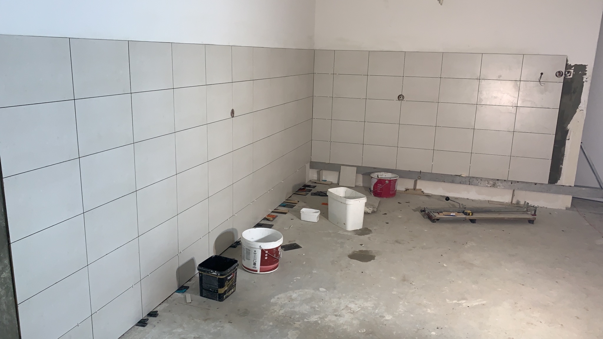 Ściana w trakcie układania białych, kwadratowych płytek. Widoczne puste puszki po farbach i narzędzia glazurnicze na podłodze.