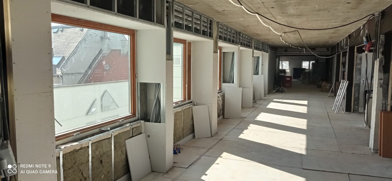 Długi korytarz w trakcie prac wykończeniowych, widoczne okna z drewnianą ramą, metalowy stelaż pod płyty gipsowe, ocieplenie z wełny mineralnej i porozkładane płyty karton-gips.