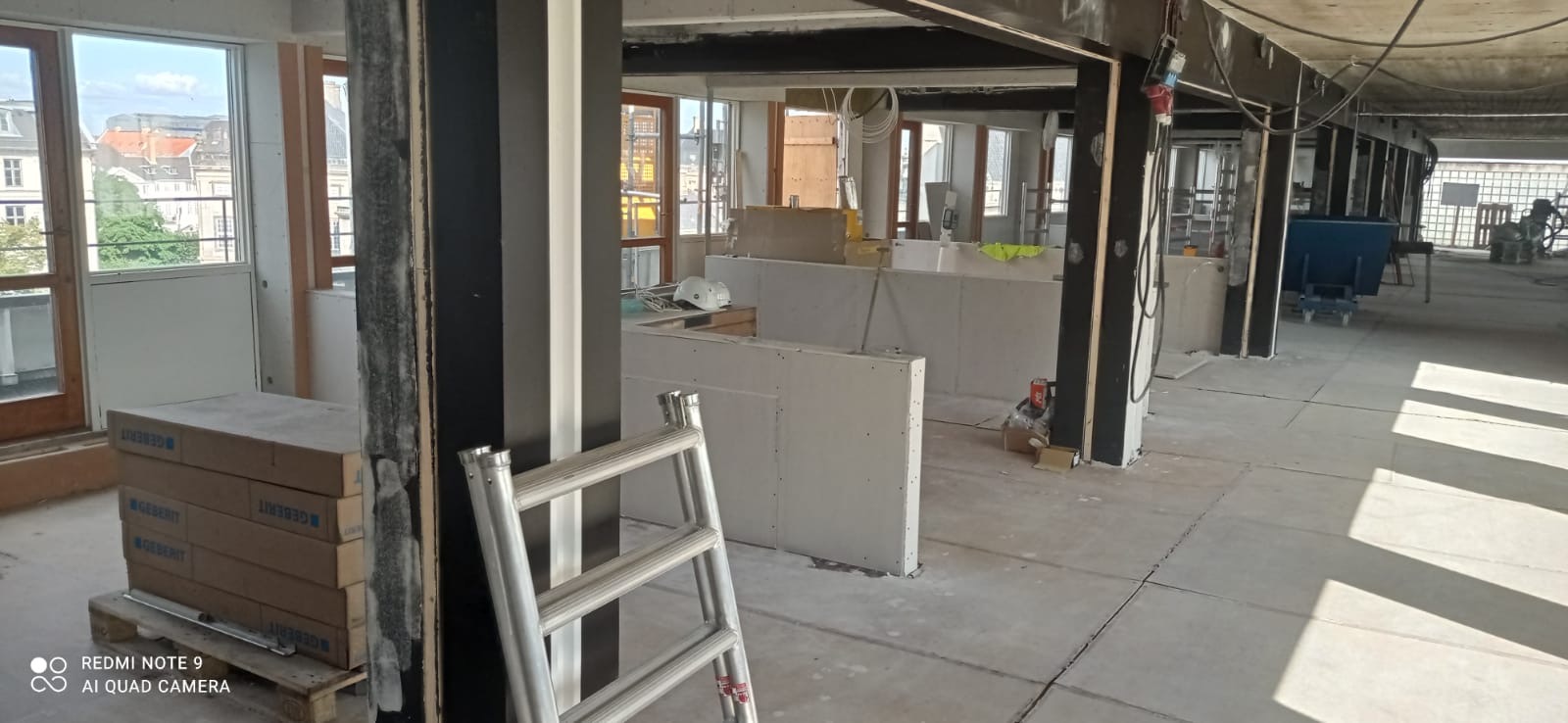 Wnętrze w trakcie prac wykończeniowych: widoczne płyty gipsowo-kartonowe, paleta z opakowaniami Geberit, aluminiowa drabina oparta o filar, biały kask ochronny i narzędzia w tle. Surowy beton...