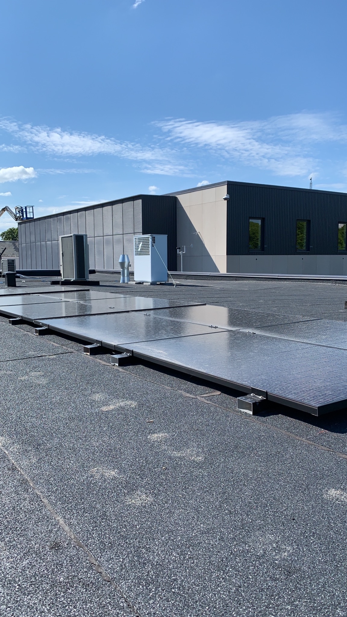 Instalacja paneli słonecznych na płaskim dachu budynku przemysłowego, widoczne panele, klimatyzatory i pracownicy w oddali, błękitne niebo.