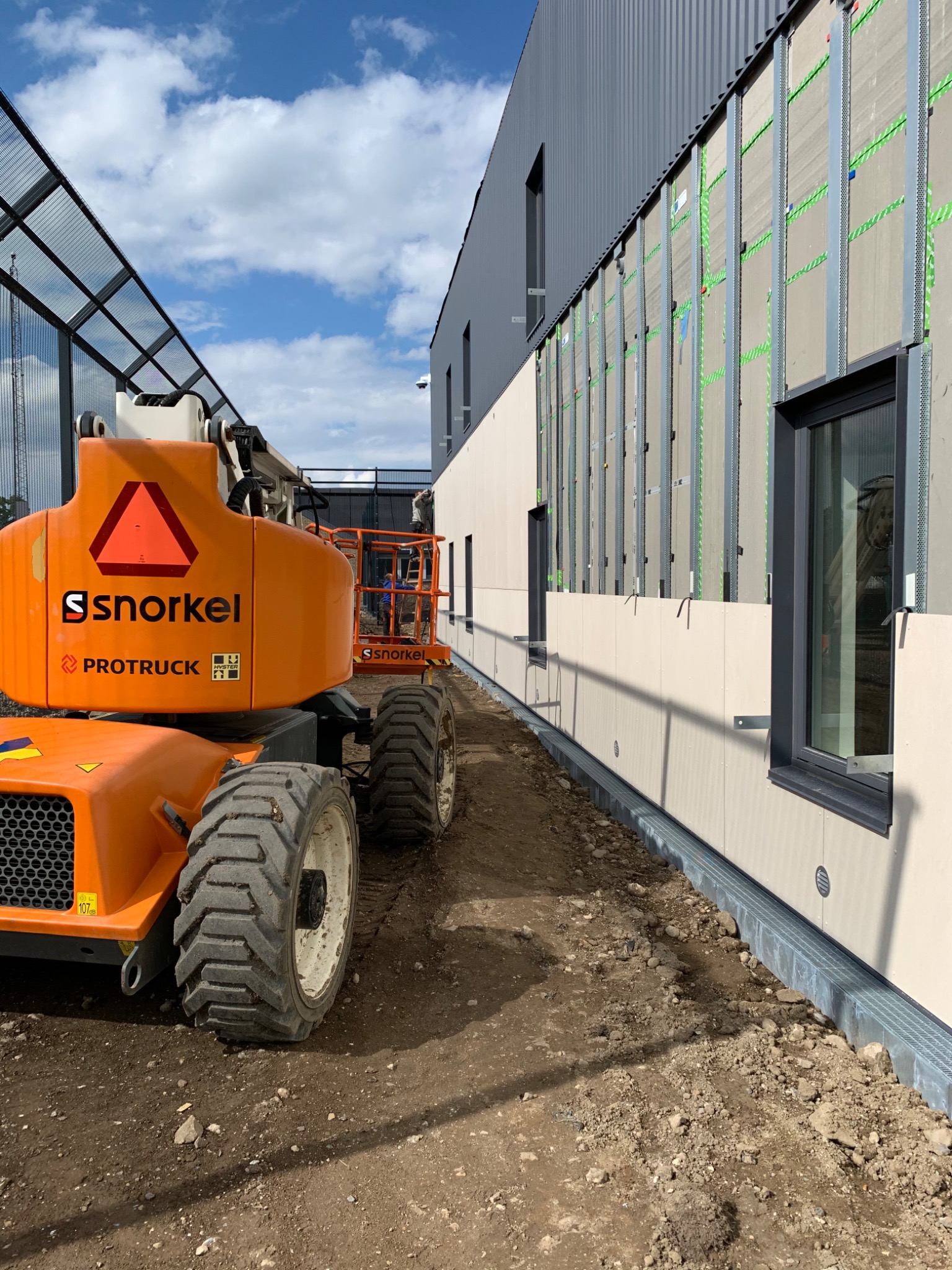 Montaż elewacji budynku z użyciem podnośnika koszowego Snorkel, widoczne profile mocujące i płyty elewacyjne w trakcie instalacji.