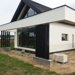 Nowoczesny dom jednorodzinny z płaskim dachem i dużymi oknami, widoczna elewacja w kolorze białym i czarne elementy konstrukcyjne, w tle zielona trawa i częściowo widoczne inne budynki.