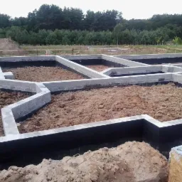 Zalane betonem fundamenty domu jednorodzinnego z hydroizolacją, widoczne zbrojenia. Tło stanowią drzewa.