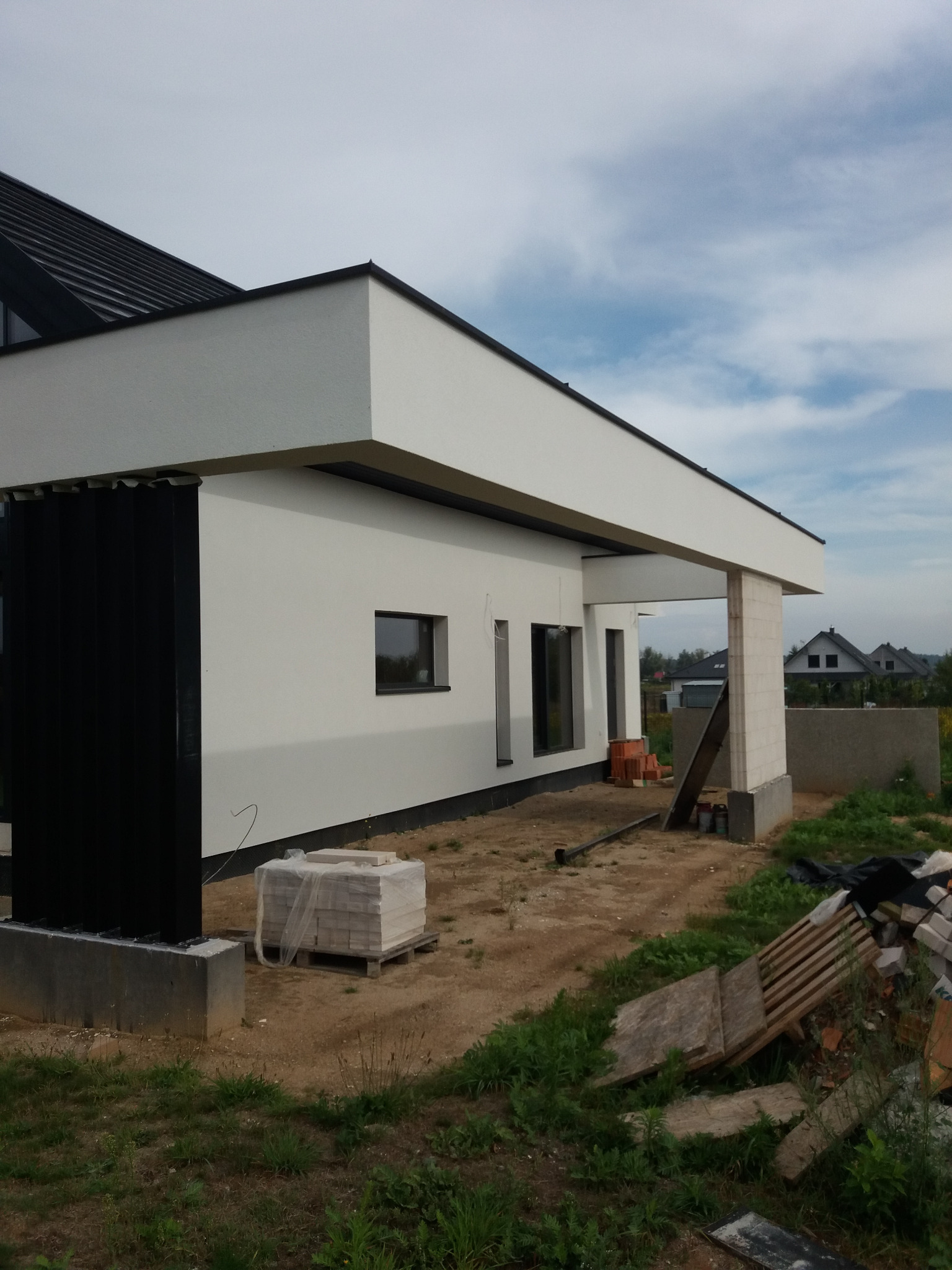 Surowy stan domu jednorodzinnego z białym tynkiem, czarnymi ramami okiennymi i elementami elewacji, widoczne materiały budowlane na placu budowy.