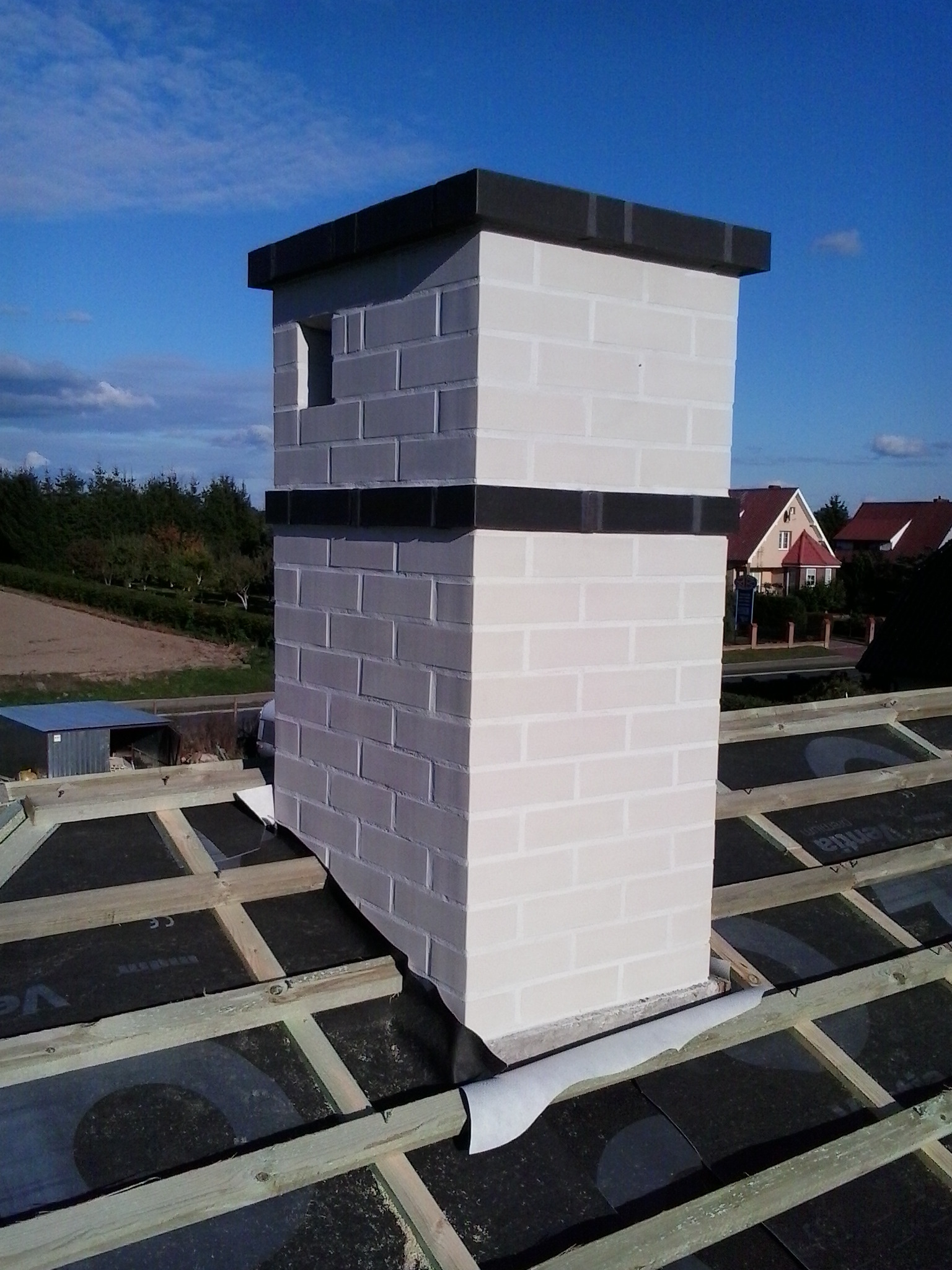 Wzniesiony z białej cegły komin z czarnymi akcentami, widoczny na tle nieba i częściowo wykończonego dachu z membraną i drewnianą konstrukcją.