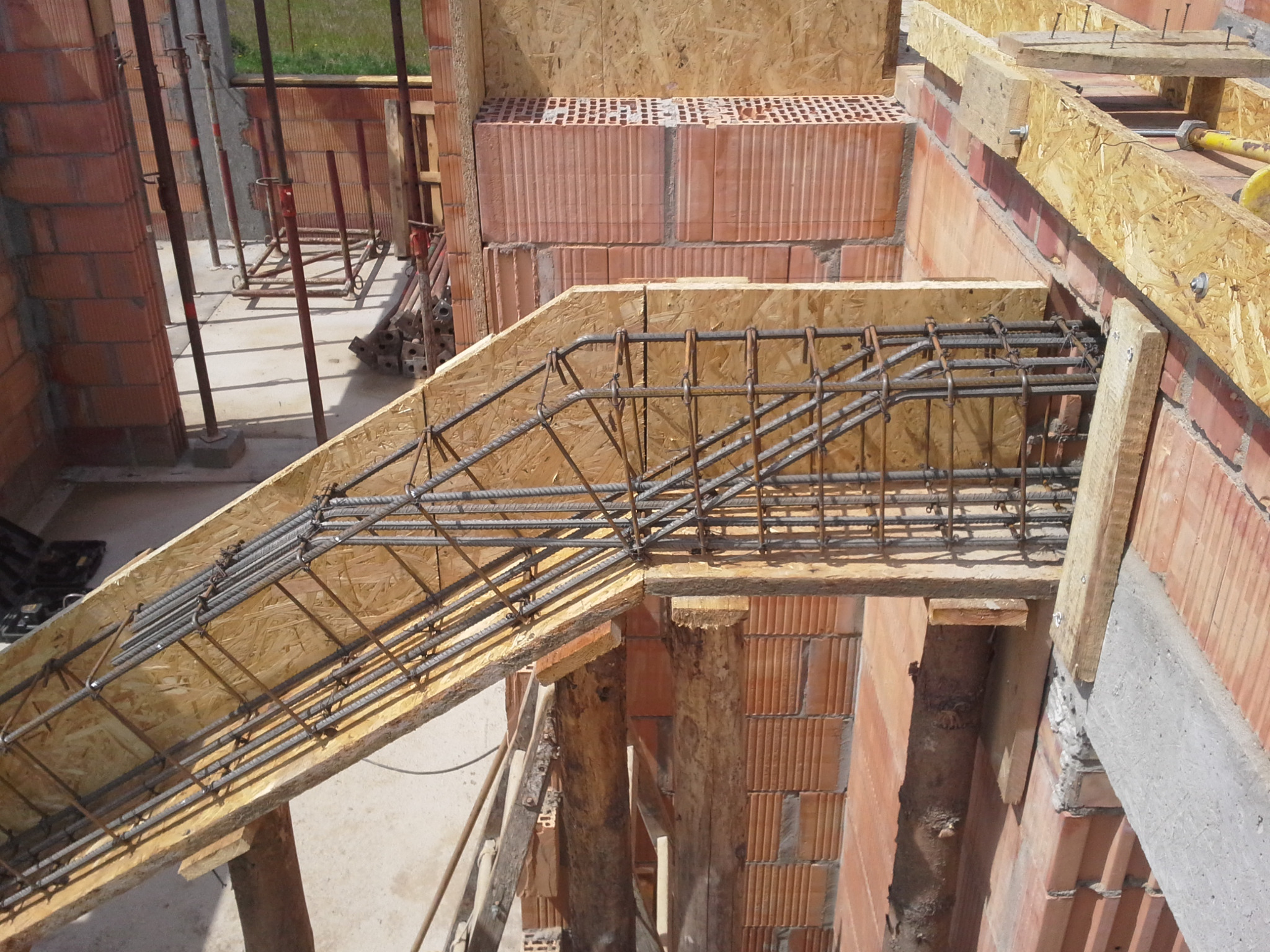 Wiązanie zbrojenia schodów w szalunku z płyty OSB, widoczne ceglane ściany domu w trakcie budowy.