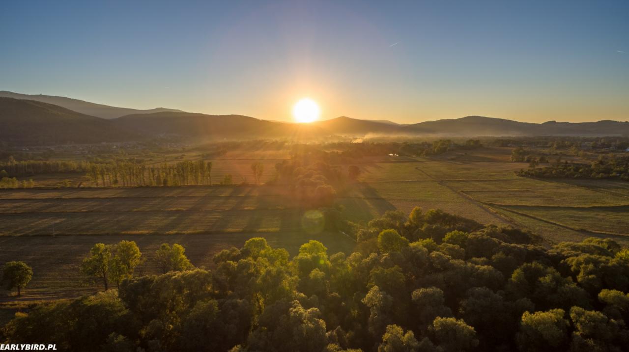 Pejzaż z lotu ptaka: wschodzące słońce nad zalesionymi wzgórzami i polami, delikatna mgła w dolinach, ciepłe światło podkreśla fakturę drzew i ziemi.