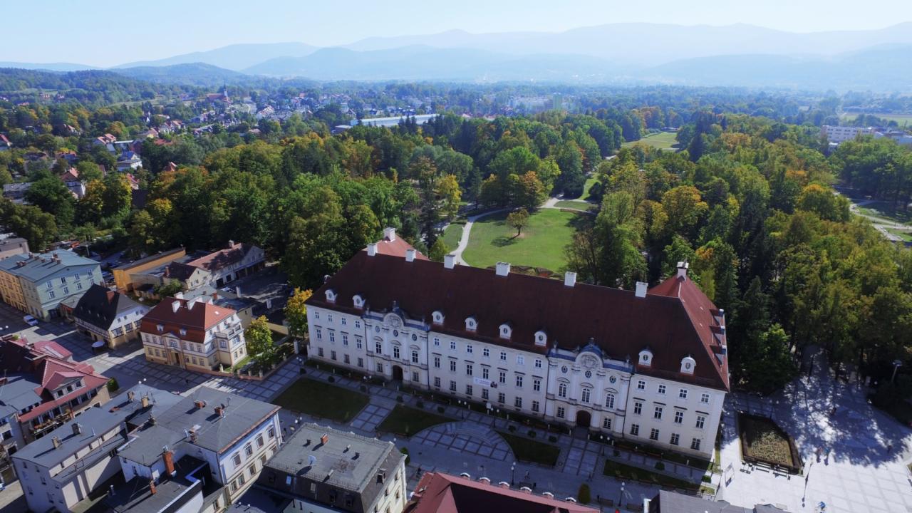 Panorama z lotu ptaka: klasycystyczny pałac z czerwoną dachówką, otoczony zielenią i zabudową miejską. W tle góry, błękitne niebo. Ujęcie z drona.
