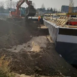 Pomarańczowa koparka z łyżką wysypuje ziemię na placu budowy, widoczni pracownicy w odblaskowych kamizelkach przy zabezpieczonym wykopie fundamentowym.