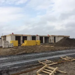 Budowa domu z betonu komórkowego w trakcie realizacji. Widoczne palety z bloczkami Ytong i robotnicy na murach. Gruntowa droga i pochmurne niebo.