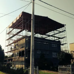 Budynek w trakcie renowacji elewacji, otoczony rusztowaniem z zabezpieczeniem. Widoczni pracownicy na rusztowaniu. Ujęcie z dołu, podkreślające wysokość budynku.