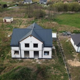 Blachotrapez - Odział Smardzów koło Oleśnicy - Niedokończony dom dwupiętrowy z ciemnoszarą blachą na dachu, widok z góry, w tle dym i inne budynki.