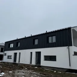 Nowoczesny dom szeregowy z czarnym dachem z blachy na rąbek stojący i białymi ścianami, widoczne rynny i kominy wentylacyjne, teren wokół budynku w trakcie prac ziemnych, resztki śniegu.