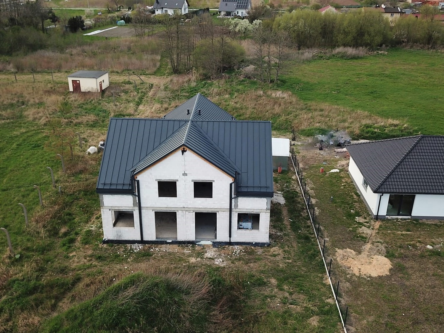 Niedokończony dom dwupiętrowy z ciemnoszarą blachą na dachu, widok z góry, w tle dym i inne budynki.