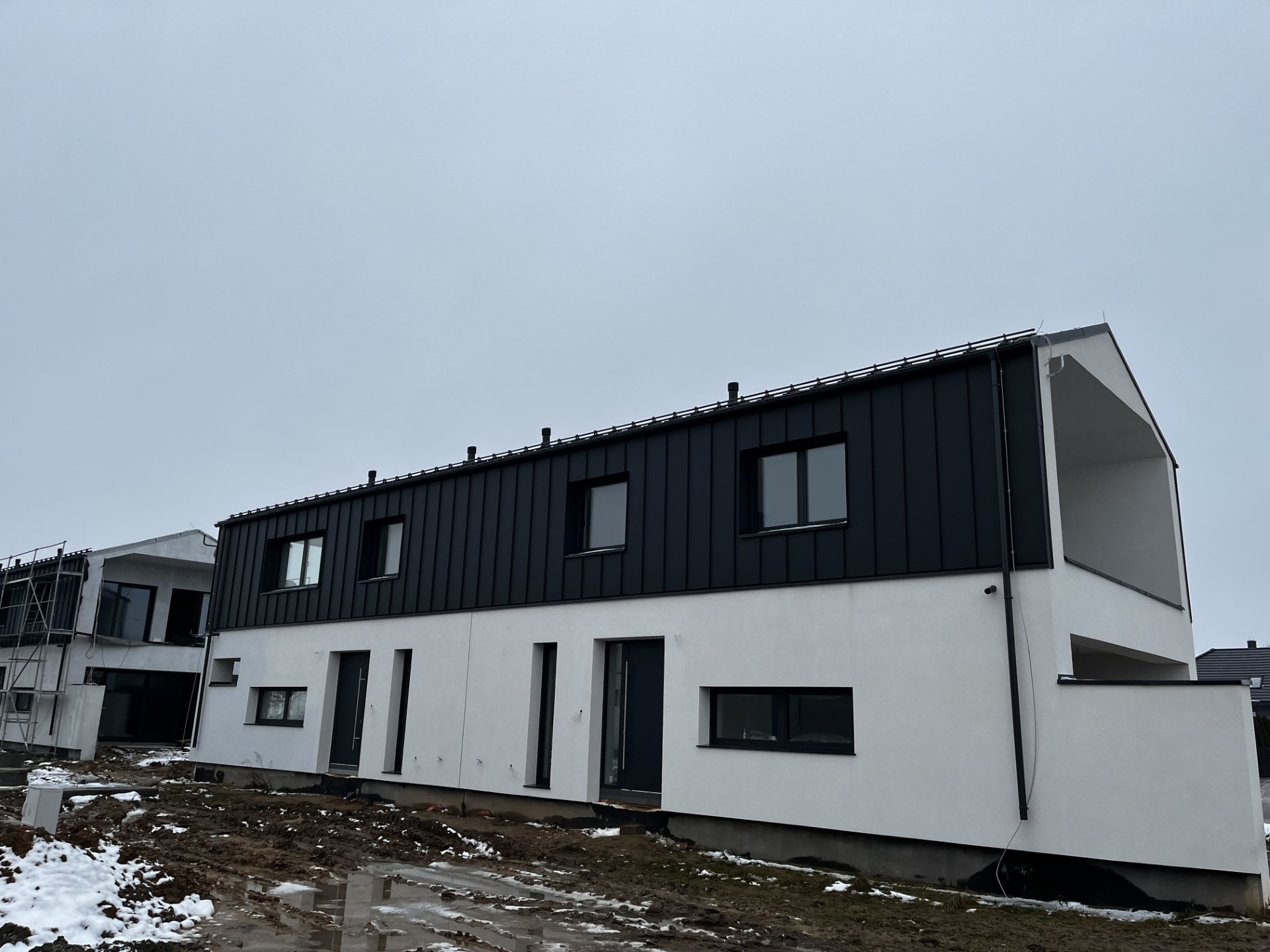 Nowoczesny dom szeregowy z czarnym dachem z blachy na rąbek stojący i białymi ścianami, widoczne rynny i kominy wentylacyjne, teren wokół budynku w trakcie prac ziemnych, resztki śniegu.