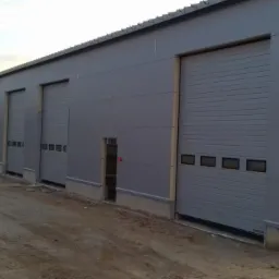 Szary budynek przemysłowy z kilkoma segmentowymi bramami garażowymi i małym wejściem po lewej stronie, widoczne ślady prac ziemnych.