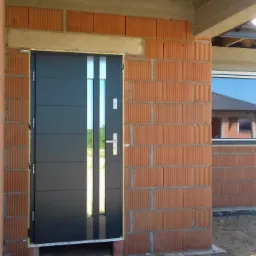 Nowoczesne, antracytowe drzwi wejściowe z dwoma pionowymi, srebrnymi paskami, zamontowane w surowym murze z czerwonej cegły dziurawki, widoczne okno z odbiciem budynku w tle.