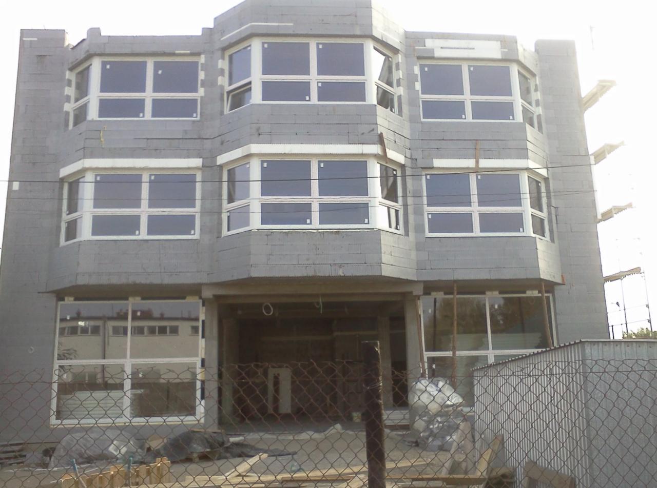 Surowy budynek w trakcie budowy z zamontowanymi oknami PCV z białymi ramami, widoczne rusztowania i materiały budowlane.