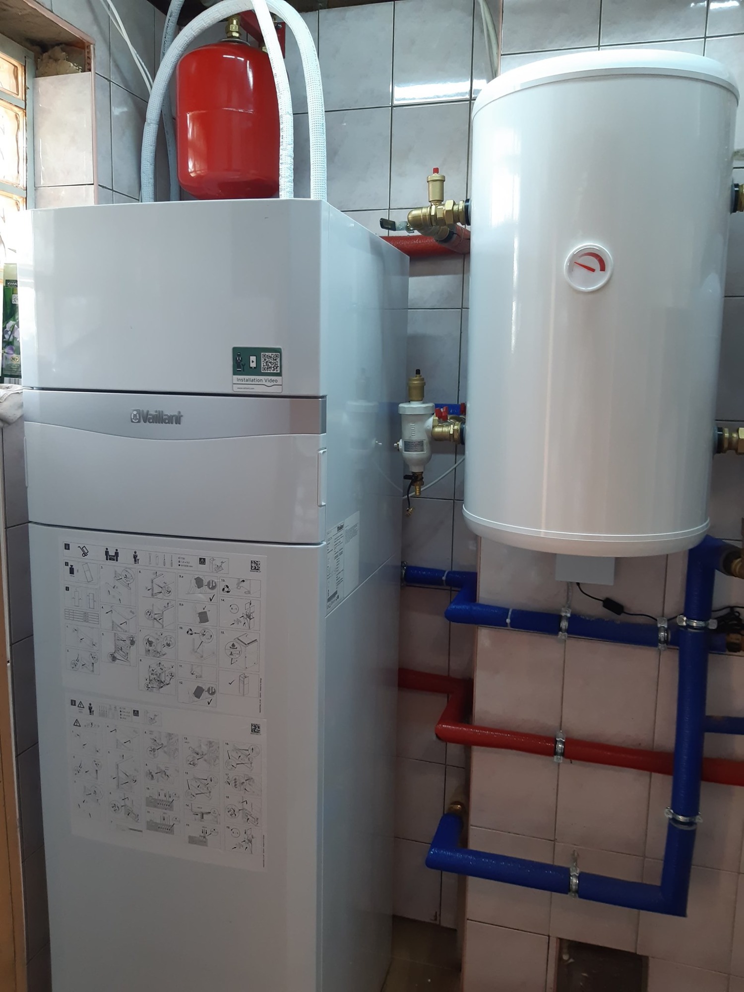 Biała pompa ciepła Vaillant z czerwonym zbiornikiem wyrównawczym i białym zasobnikiem ciepłej wody, podłączona do instalacji z rurami w kolorze niebieskim i czerwonym, zamontowana w pomieszczeniu...