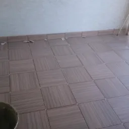 Układanie brązowych płytek imitujących drewno na podłodze balkonu, widoczny kubeł z zaprawą, fragment balustrady i resztki po cięciu płytek.