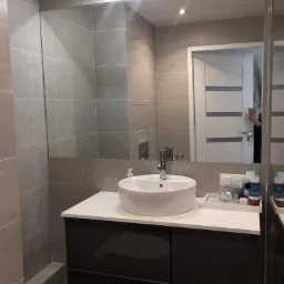 Nowoczesna łazienka z szarymi płytkami, okrągłą umywalką nablatową i ciemnoszarą szafką pod umywalką; w lustrze odbicie fragmentu białych drzwi.