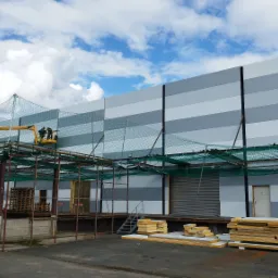 Montaż płyt warstwowych na elewacji magazynu z użyciem podnośnika koszowego i zabezpieczeń siatkowych, widoczne przygotowane płyty i konstrukcja stalowa.