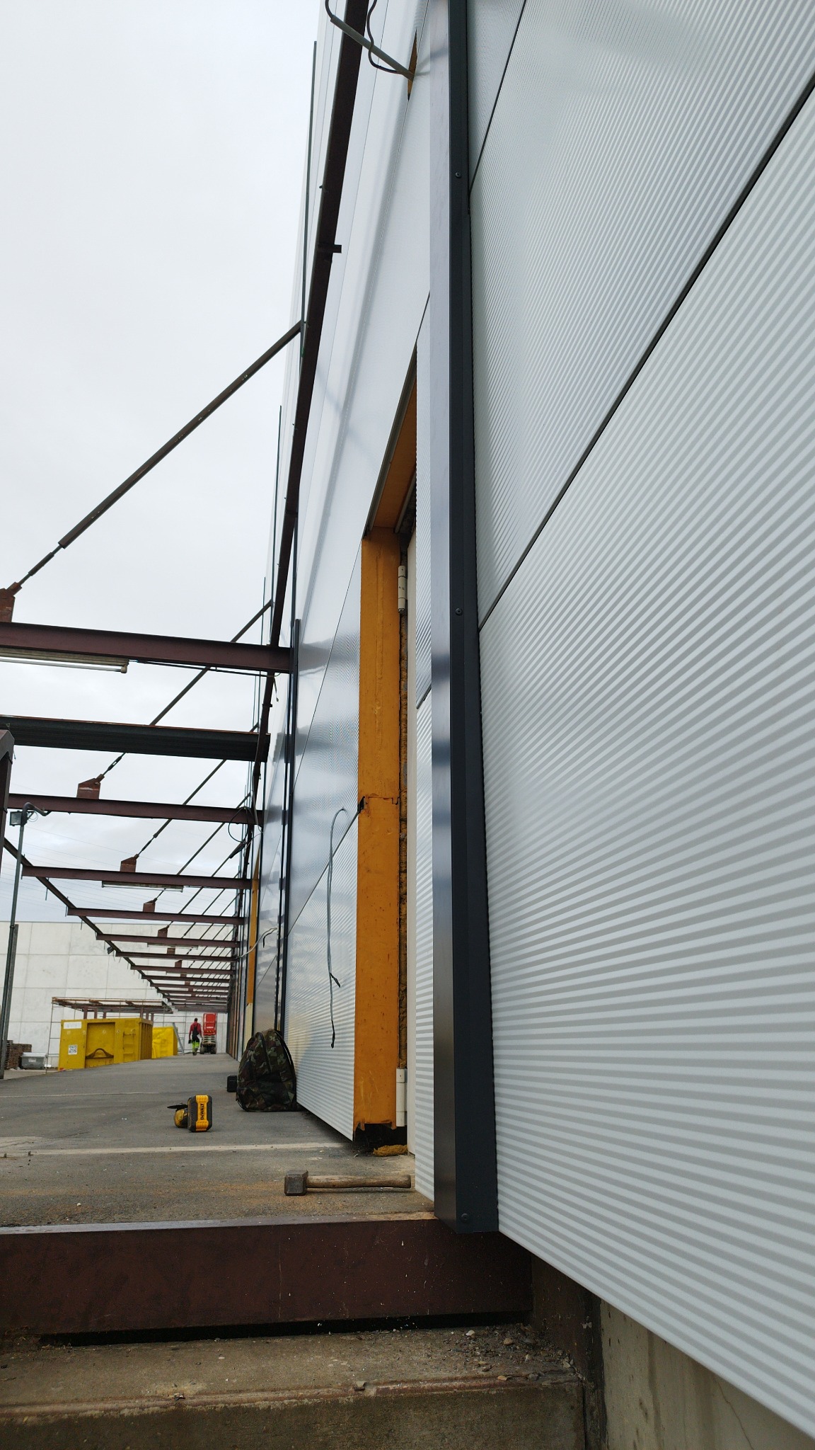 Montaż szarej płyty warstwowej z widocznym fragmentem ramy okiennej, narzędzia pracy na ziemi, w tle konstrukcja stalowa i osoba w oddali.