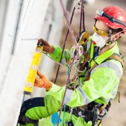 Pracownik w kasku i uprzęży, zabezpieczony linami, poziomuje ścianę budynku w Poznaniu za pomocą poziomicy. Widoczne narzędzia i osprzęt do prac na wysokości.