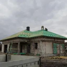 Budowa domu murowanego: Dwóch pracowników na dachu pokrytym zieloną membraną, montujących elementy konstrukcyjne, widoczne rusztowania i częściowo otynkowane ściany.