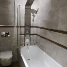 Wykończona łazienka z szarymi płytkami, wanną z panelem prysznicowym, toaletą podwieszaną i blatem z jasnego drewna.