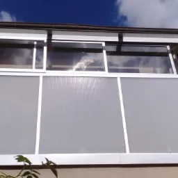 Zabudowany balkon z aluminiową ramą i półprzezroczystym wypełnieniem, widoczna antena satelitarna na elewacji budynku.