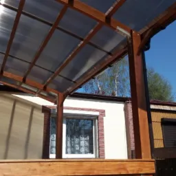 Zadaszenie tarasu z drewnianą konstrukcją i przezroczystym poliwęglanem, widok z dołu na tle budynków w Wągrowcu.