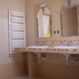 Jasna łazienka z dwiema umywalkami, dużymi lustrami z oświetleniem oraz grzejnikiem drabinkowym na ścianie, widoczne beżowe płytki i drewniane drzwi.