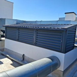 ALU-STAL - Szara osłona wentylacyjna z żaluzjami na dachu budynku, widoczne rury i elementy instalacji, błękitne niebo w tle.