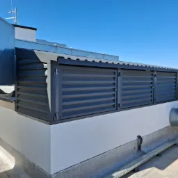 Szare, poziome panele osłonowe na dachu budynku, z widocznymi elementami wentylacji i anteną na tle błękitnego nieba.