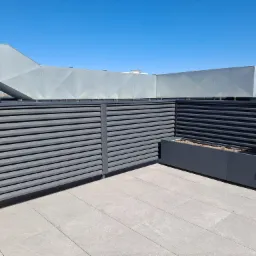 Szare poziome panele ogrodzeniowe na tle błękitnego nieba i zabudowań miejskich, z donicą na rośliny w rogu.