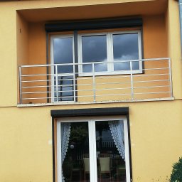 ALU-STAL - Żółta elewacja domu z balkonem zabezpieczonym prostą, poziomą, stalową konstrukcją, okna z roletami i firankami.