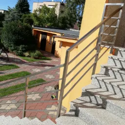 Zewnętrzne schody granitowe z balustradą ze stali nierdzewnej, prowadzące do wejścia budynku o żółtej elewacji, widok z góry na ozdobny chodnik z kostki i trawy w tle.