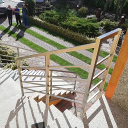 ALU-STAL - Zewnętrzne schody z jasnego kamienia z nowoczesną, prostą balustradą ze stali nierdzewnej, widok z góry na podjazd z kostki i trawy oraz grupę osób przed domem.